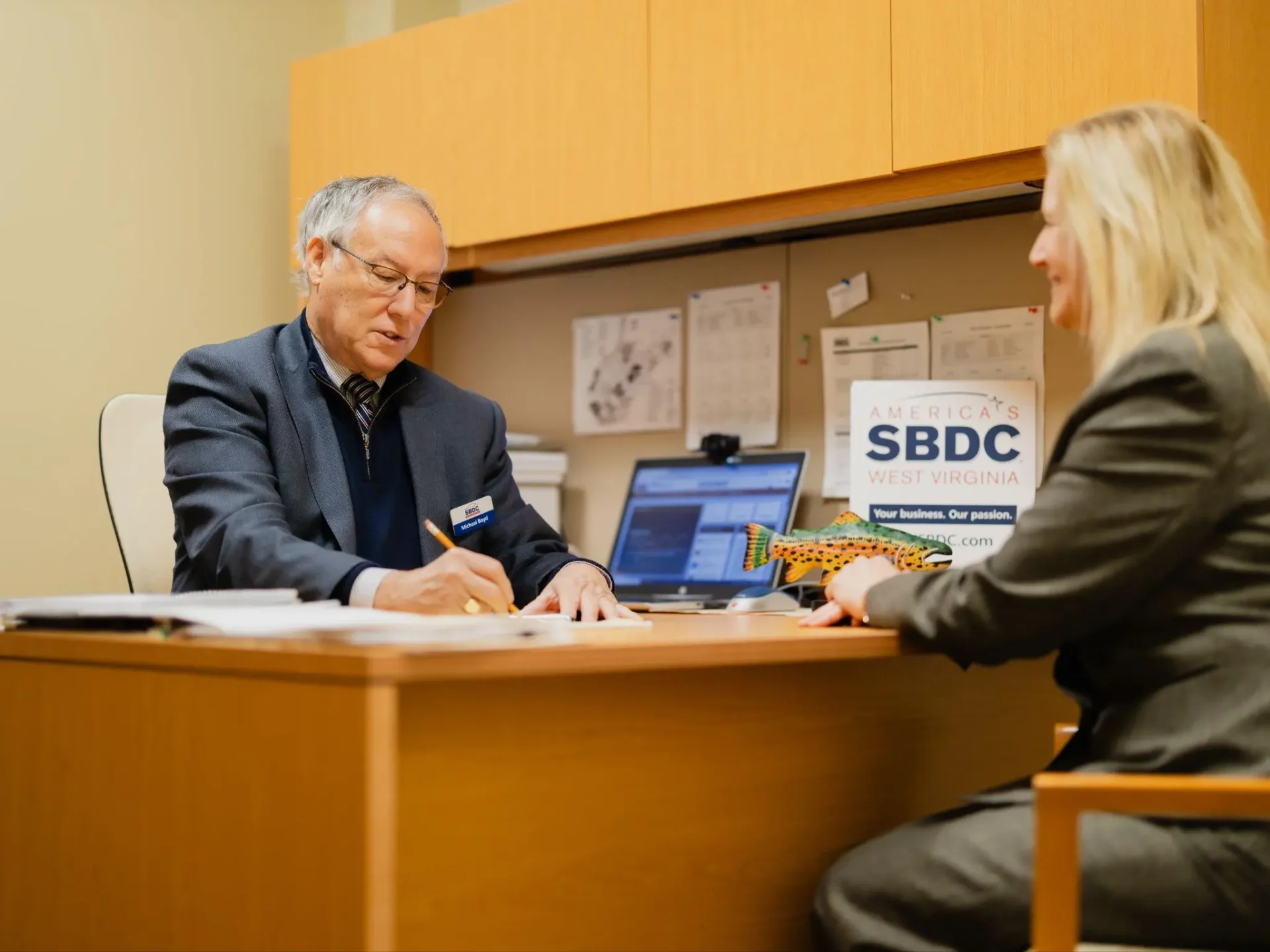 Two people are seated in an office, having a discussion. One person is taking notes, while the other is holding a model fish. A sign for SBDC West Virginia is visible on the desk.