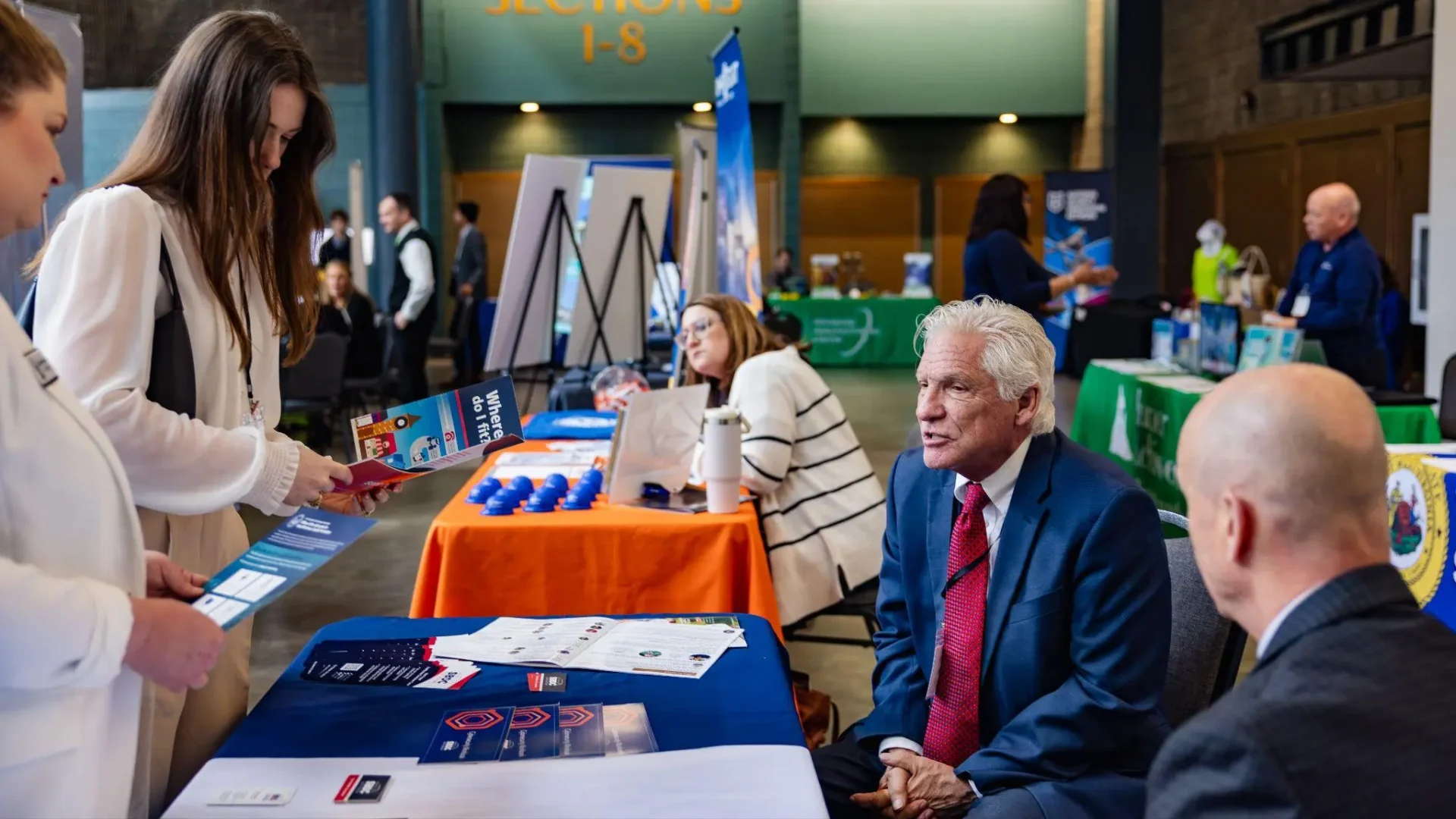 
People are interacting at a business expo. Two women are looking at brochures at a booth, while two men are seated and engaged in conversation. Various informational booths and banners are visible in the background.