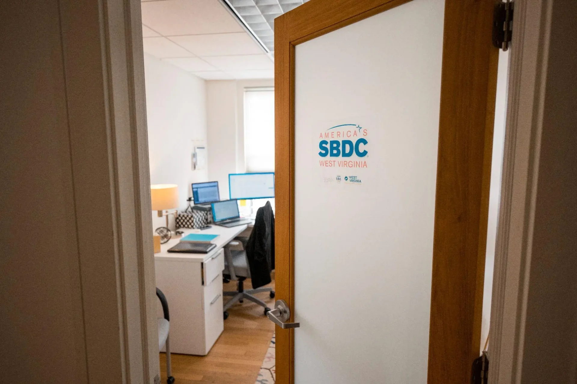 An office space is shown through a partially open door, featuring multiple computer monitors, a desk with office supplies, a chair, and a lamp. The door has a sign for SBDC West Virginia.